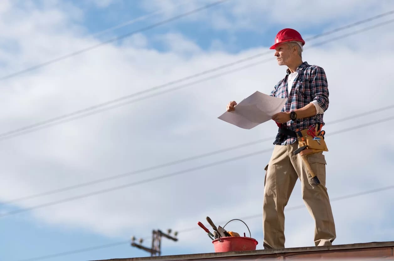 A roofer stands on a roof inspecting construction blueprints
