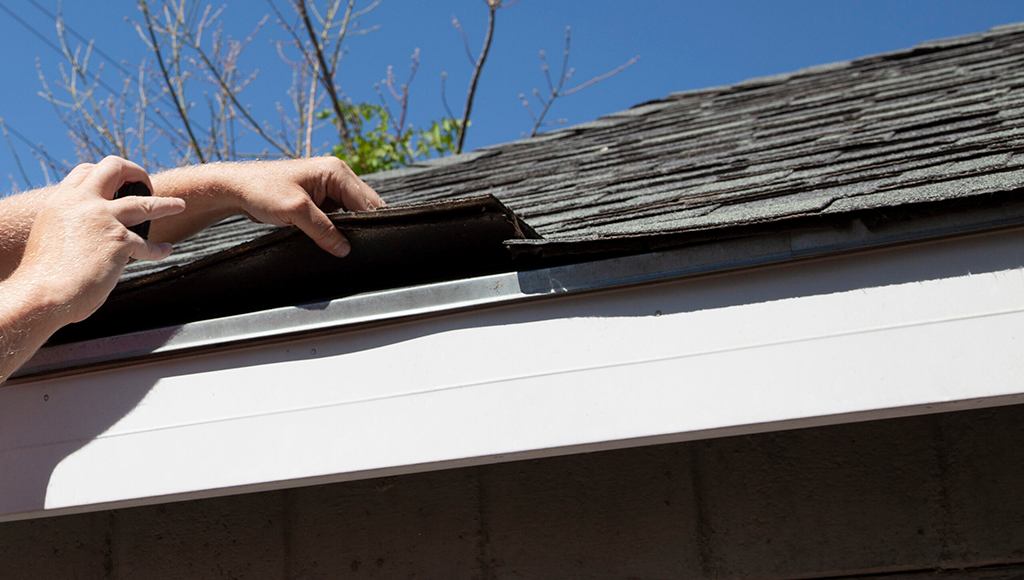 A person lifts up the edges of shingles on an asphalt shingle roof