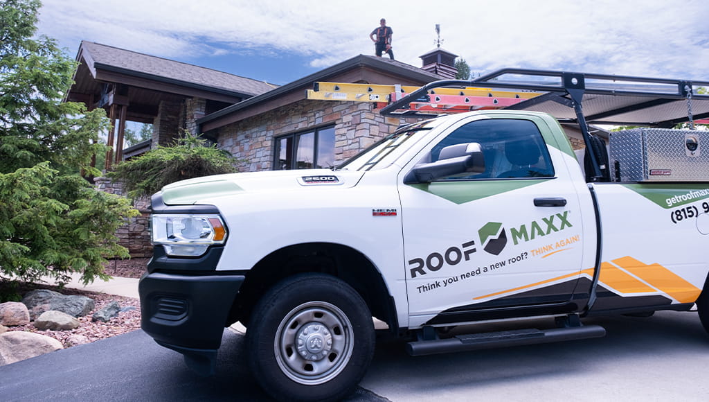 Roof Maxx work truck parked outside of a residence where a worker is standing on the roof.