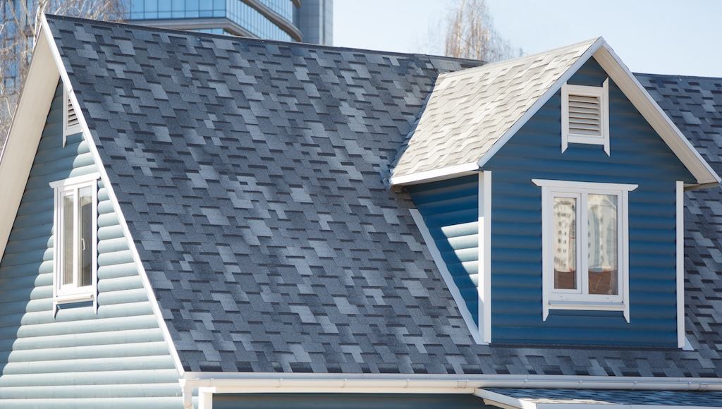 A tall peaked roof with a gable feature on a grey residential home with asphalt shingles.