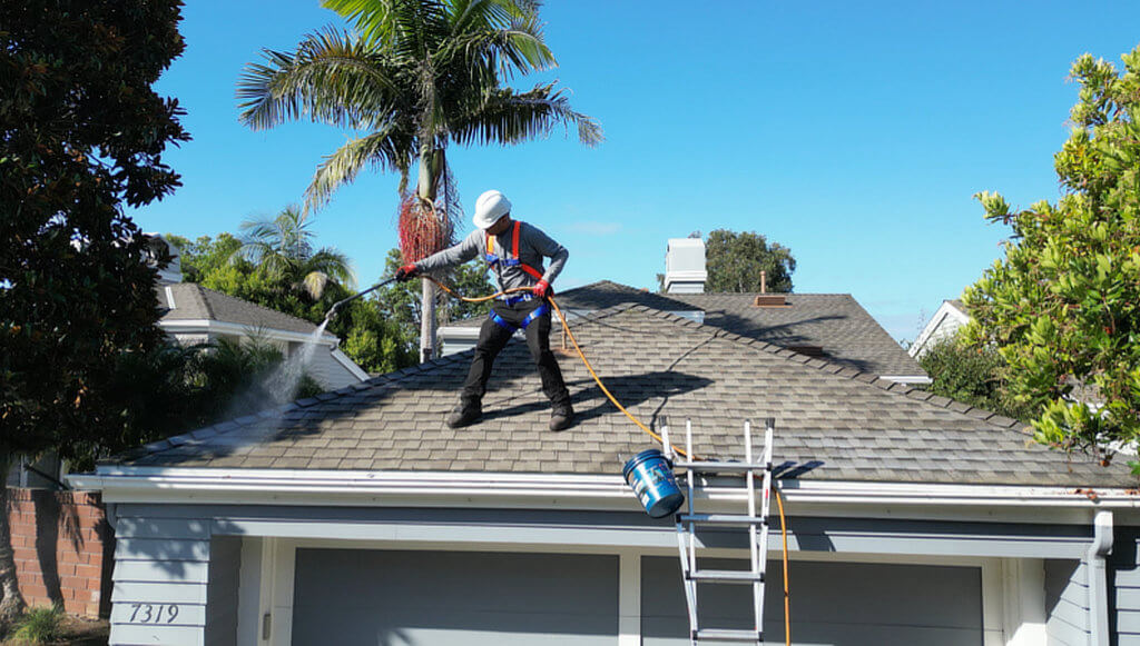 A Roof Maxx dealer sprays treatment on a residential roof against a clear blue sky background.