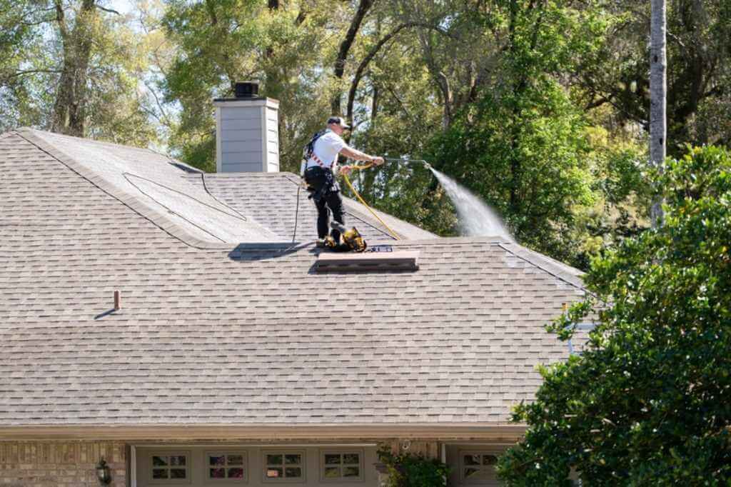 A Roof Maxx professional sprays treatment solution onto asphalt shingles from a residential rooftop.