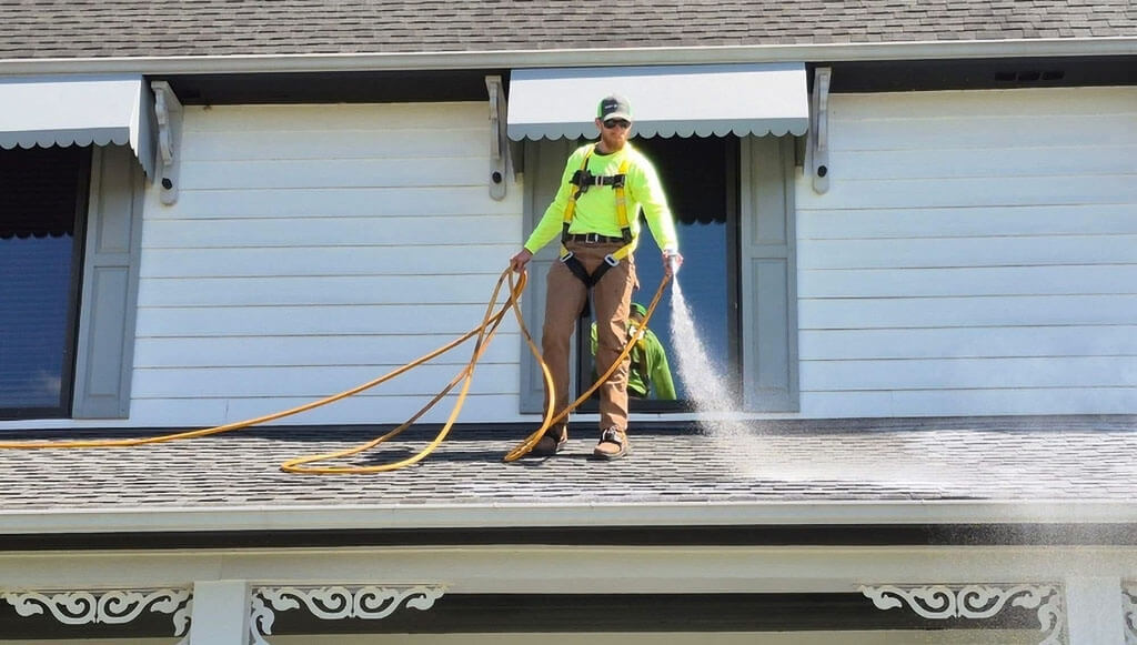 A Roof Maxx dealer sprays the treatment onto a beige asphalt shingle roof with wooden filigree accents.