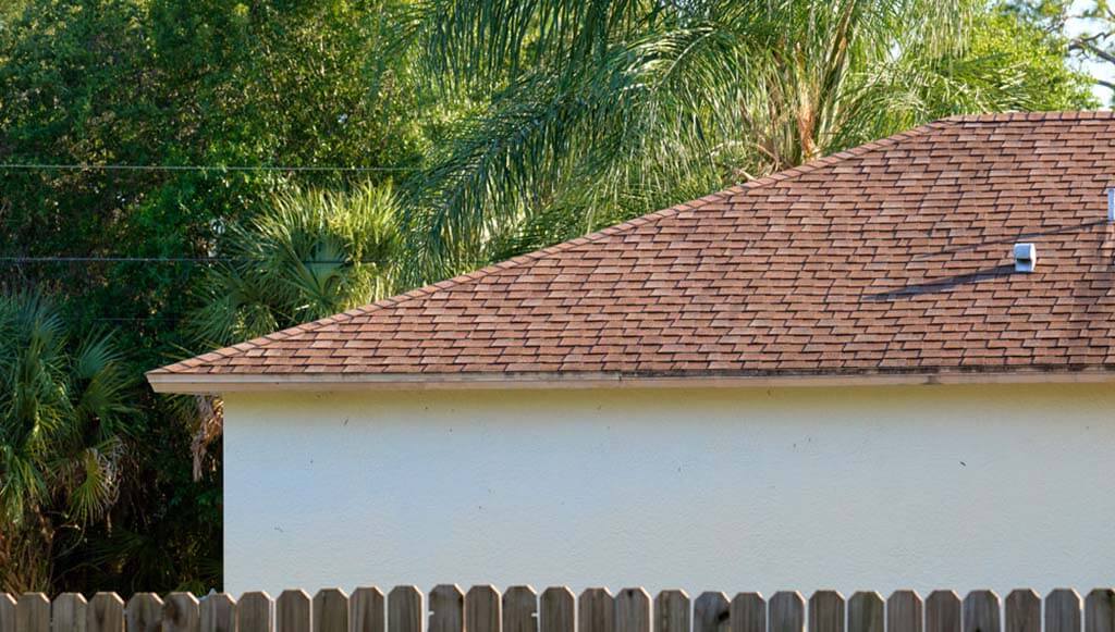 A red asphalt shingle roof on a stucco home surrounded by tropical trees.