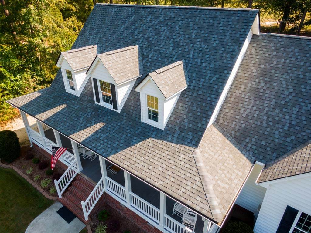 Aerial drone view of a large residential home with a multi-pitched asphalt shingle roof surrounded by trees.