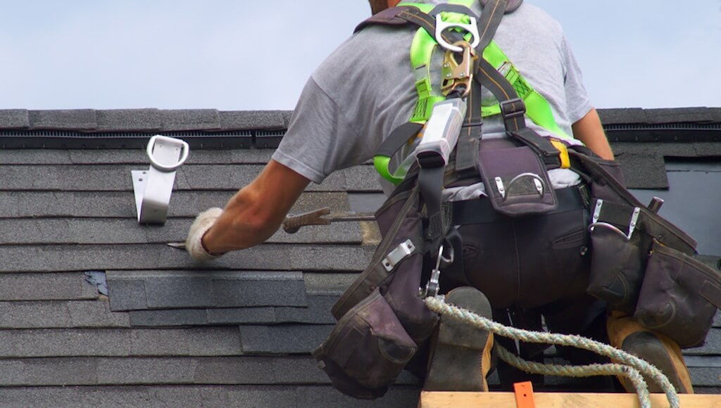 A Roof Maxx dealer wearing safety harness equipment nails shingles onto an asphalt roof.