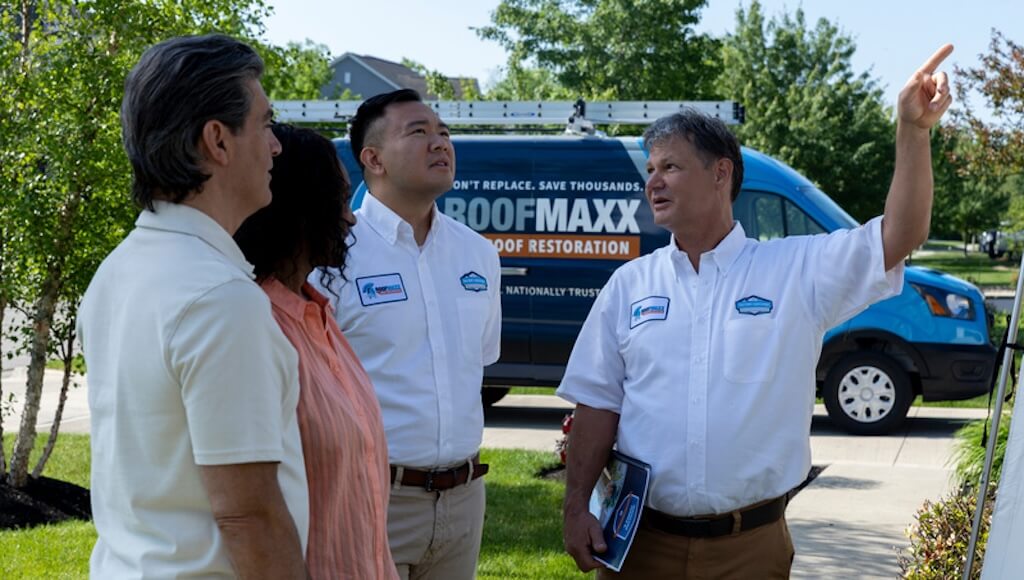 Two Roof Maxx dealers in branded shirts consult with a male and female homeowner outside their home in front of a Roof Maxx service van.