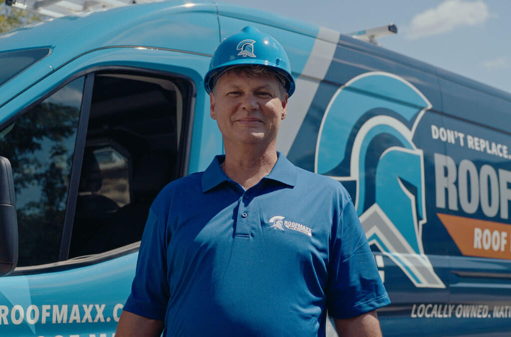 A smiling Roof Maxx dealer wearing a blue hard hat and polo shirt stands in front of a branded company van.