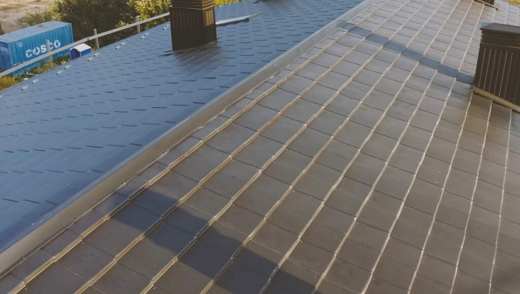 Angled aerial view of a dark metal tile gable roof with chimneys and a visible valley.