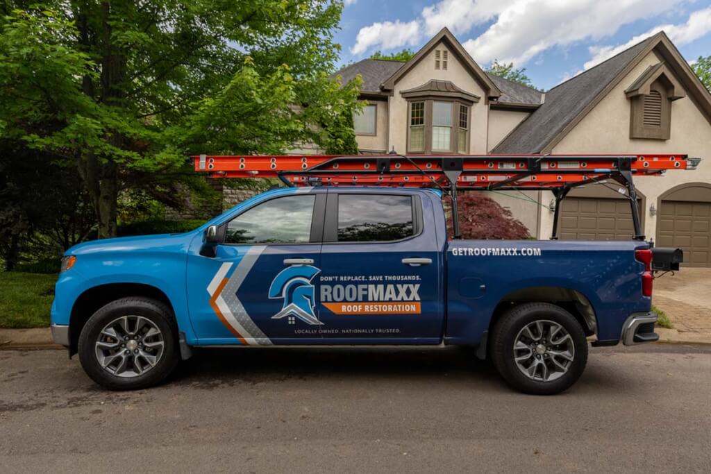A blue, grey, orange, and white branded Roof Maxx pickup truck carrying a red ladder