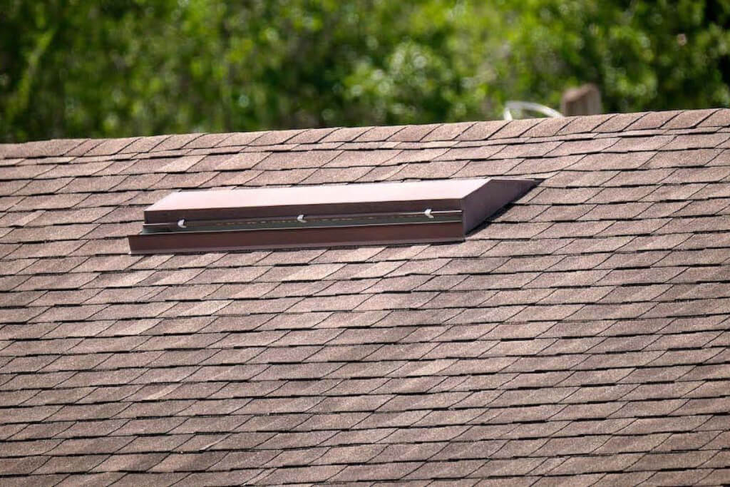 Distant shot of a brown asphalt shingle roof with a long, thin brown roof vent installed on its surface.