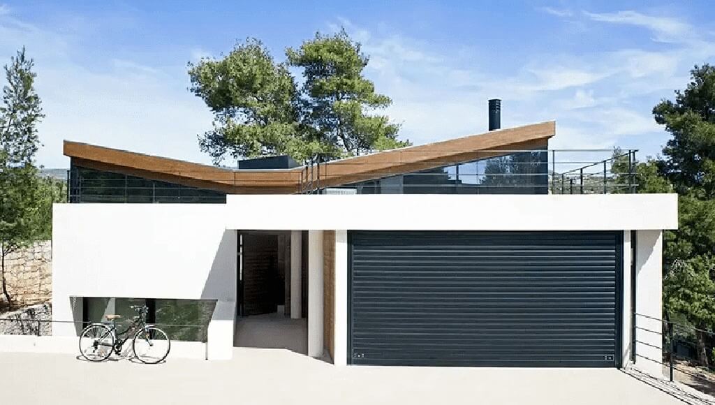 A modern home with a wooden butterfly roof, white walls, and a large dark garage door with a bicycle parked outside.