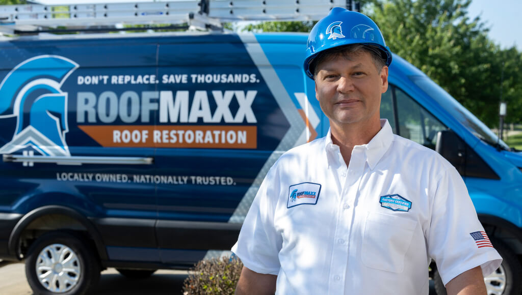 A Roof Maxx dealer wearing a blue hard hat and white uniform shirt smiles in front of his blue branded van.