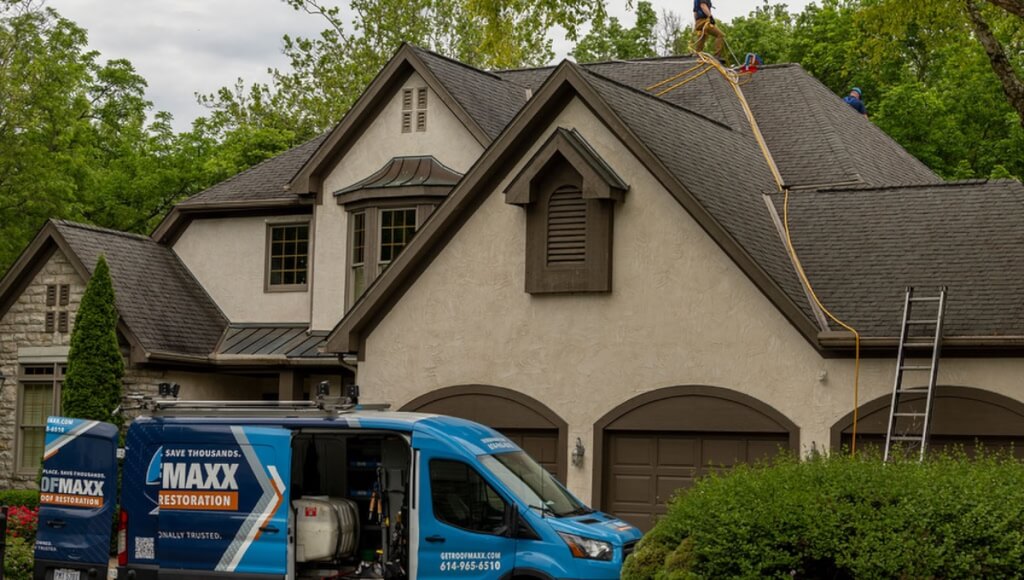 Two Roof Maxx dealers stand on a customer's asphalt shingle roof while a van is parked in the driveway below.