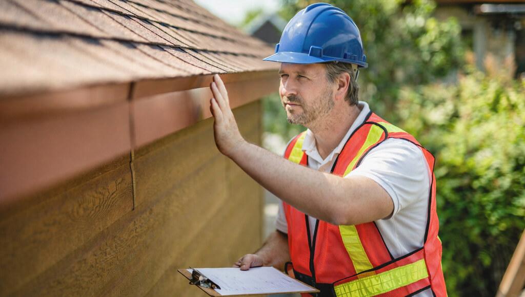 A roofer inspects the edge of an asphalt shingle roof up close.