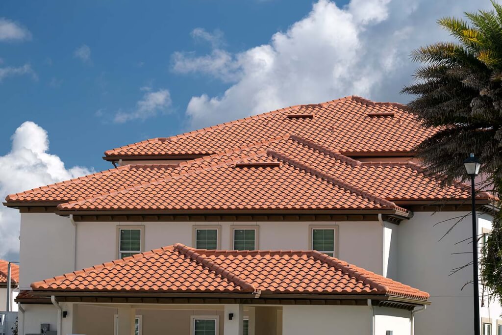A large residential home near the coast features orange clay tile roofing under a blue sky with palm trees nearby.