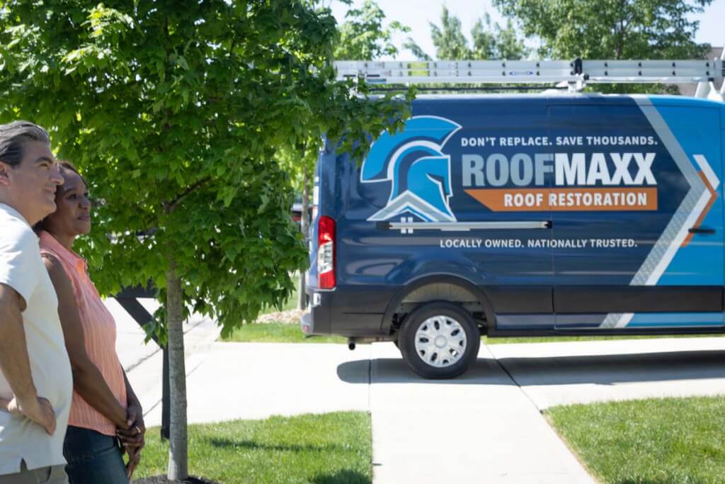 Happy homeowners stand next to a Roof Maxx branded van in a residential driveway.