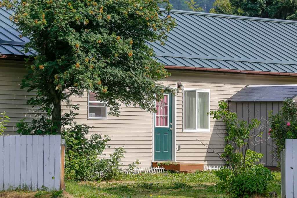 A small one-story home with aluminum panel metal roofing and a teal door surrounded by a garden and fence.