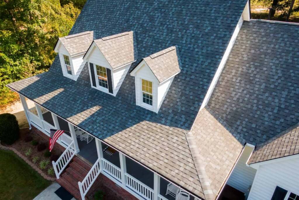 Aerial drone view of a large residential home with an asphalt shingle roof, dormers, and a covered front porch with an American flag.