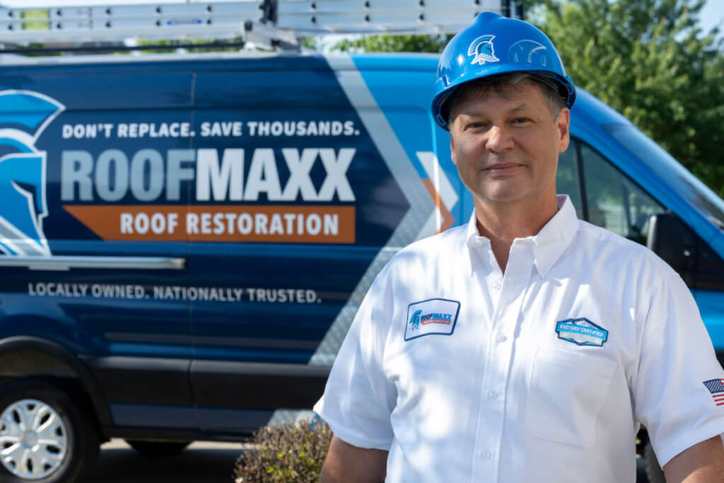 A Roof Maxx dealer wearing a blue hard hat smiles in front of his blue branded Roof Maxx van.