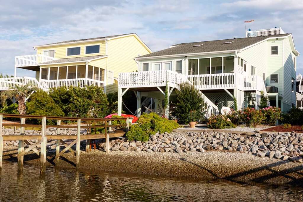 Colorful coastal homes with white decks and railings sit along a rocky shoreline near the water.