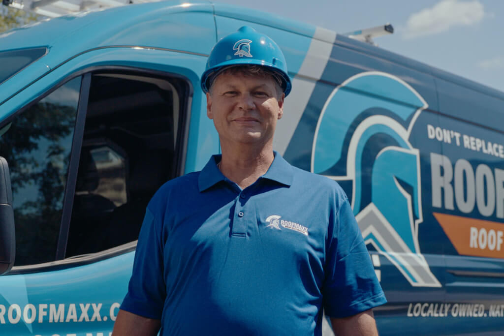A smiling Roof Maxx dealer wearing a blue hard hat and branded polo shirt stands in front of a Roof Maxx branded van.