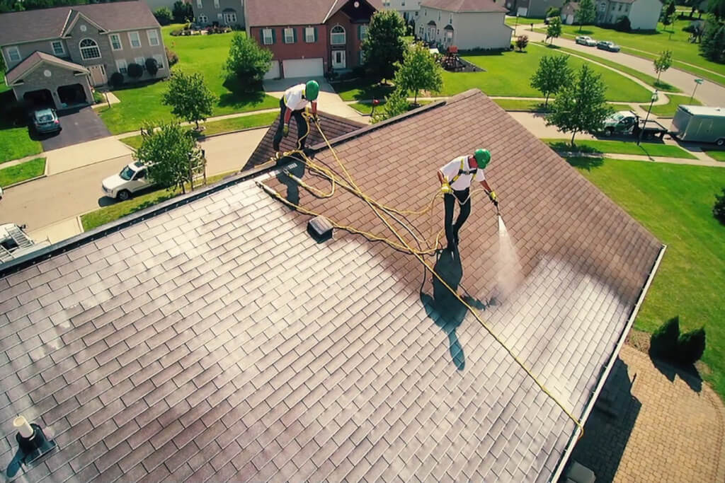Two Roof Maxx dealers in green hard hats spray an asphalt shingle roof on a sunny day in a residential neighborhood.