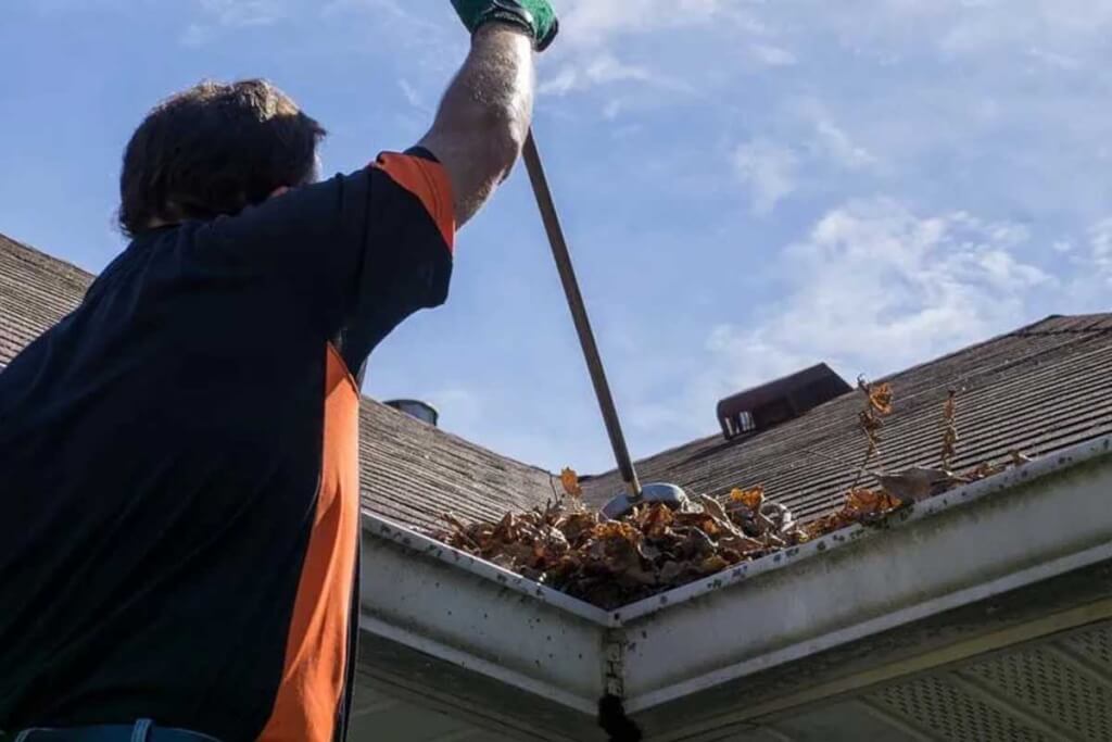 A person uses a long-handled tool to clear leaves and debris from a residential roof.