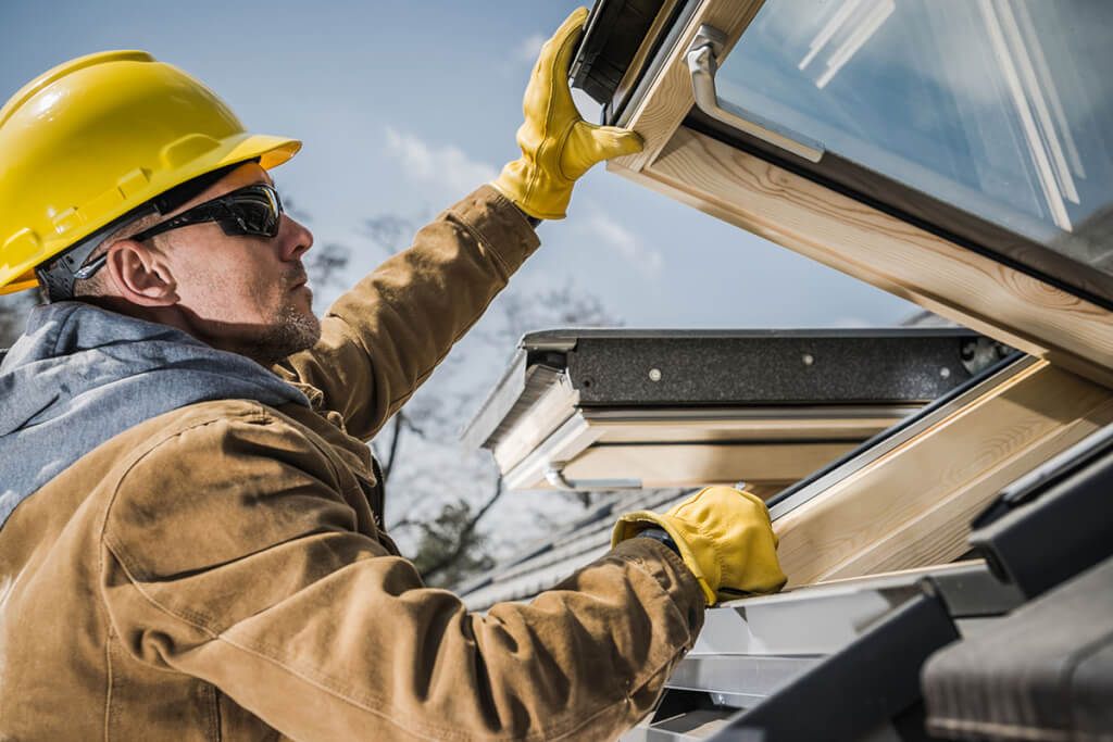A contractor closely inspects new skylights installed on an asphalt shingle roof.
