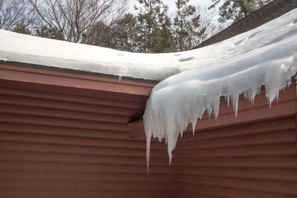 Thick ice and icicles build up along the edge of a snow-covered residential roof.