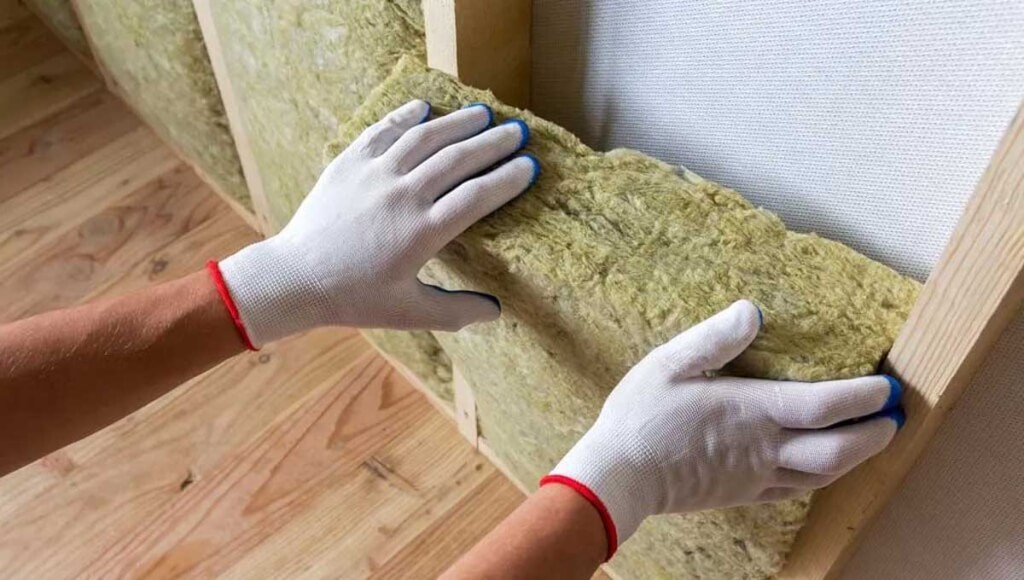A person wearing gloves installs insulation between wooden framing inside a home.