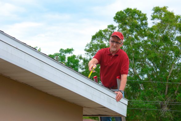 Man in red hat and shirt on top of ladder inspecting roof.