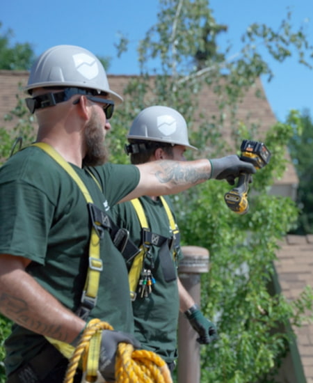 Two roofers stand on an asphalt shingle roof pointing out problem areas during a roof inspection.